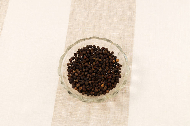 Черный перец на круглой стеклянной тарелке на полосатой льняной скатерти
. - Фото, изображение