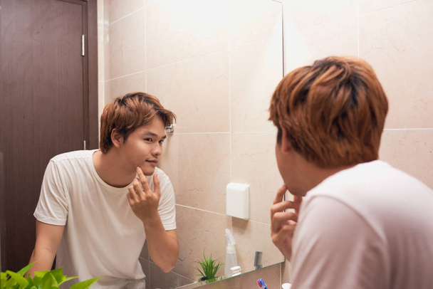 Young asian man looking in mirror and checking his skin in bathroom - Foto, Bild