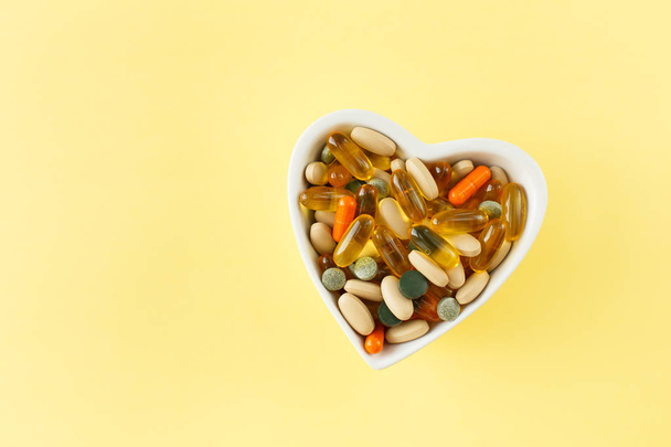 The concept of preventive health, Cup-heart with a bunch of different pills and vitamins and the omega, on a yellow background, top view - Фото, изображение