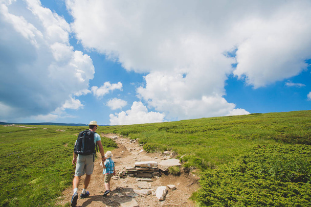 Father and son hiking in Rila mountains, Bulgaria. A traveler and a little boy are walking along the road holding hands. back view. huge white clouds on a blue sky - 写真・画像