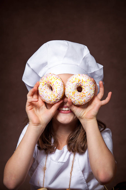 Cute girl holding two delicious donuts in her hands. Fun posing, playing in the chef's hat - Photo, Image