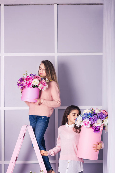 Two beautiful girls in jeans and pink sweater in studio with decor of flowers in baskets. - Zdjęcie, obraz
