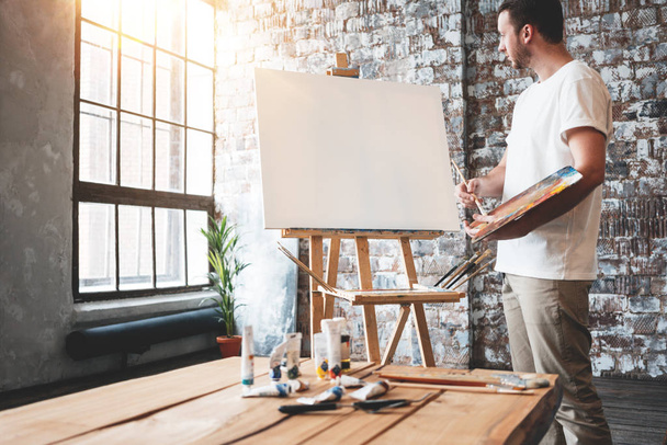 Man artist stands in front of a blank canvas on easel with palette in loft art studio. Male painter hold paintbrush in hand and mix paint. Hobby concept - Fotoğraf, Görsel