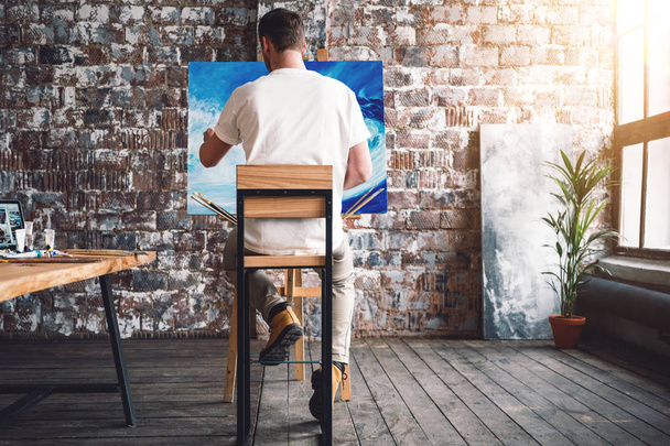 Male painter sits on chair in front canvas and drawing picture in studio. Art class and workshop. Artist painting process. Hobby - Foto, afbeelding