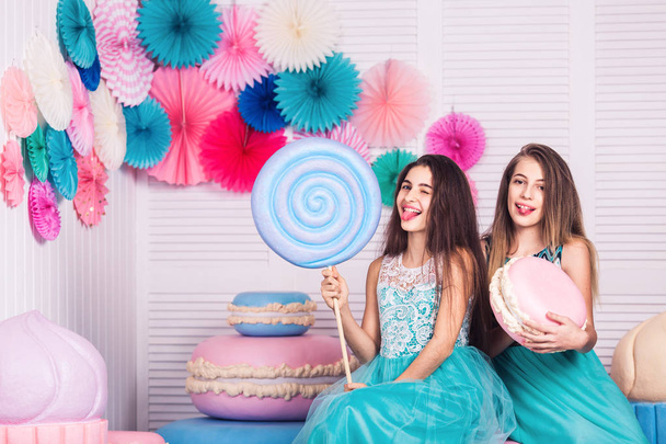 Дві красиві дівчата в блакитних сукнях тримають в руках величезні цукерки і макаруни в студії з декором макарунів
 - Фото, зображення