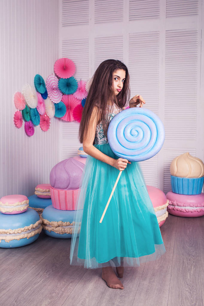 beautiful girl in blue dress hold in hand huge candy in studio with decor of macaroons. Copy space. - Foto, Imagem