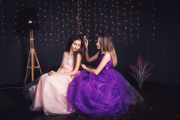 Cheerful girlfriends. Two girls comb each other's hair, sitting side by side in studio on dark background. Copy space - Photo, Image