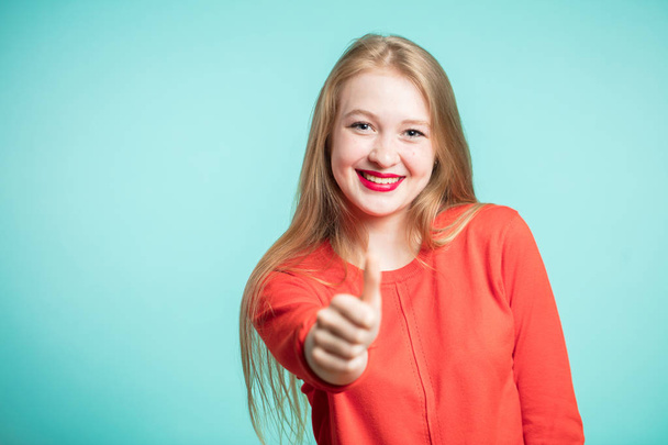 Smiling young woman in red dress shows the thumbs up on a blue background - Foto, Imagem