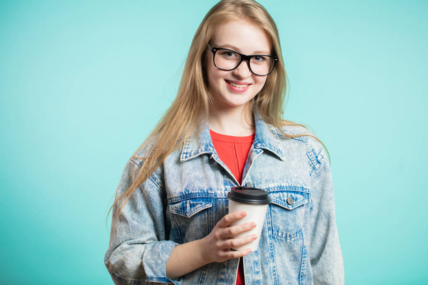 Young cute smiling girl in glasses and jeans holds coffee on his hand and looks at the camera with a smile - 写真・画像