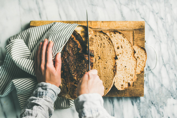Female hands cutting freshly baked sourdough bread into pieces on rustic wooden chopping board over light grey marble background - 写真・画像