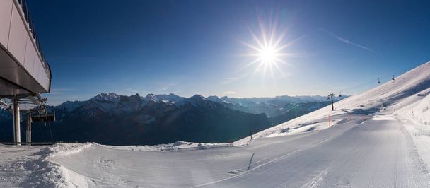 vista panorámica de una estación de esquí con pistas de esquí recién arregladas y telesillas en un hermoso día de invierno con un cielo azul y el sol brillante por encima y una gran vista detrás
 - Foto, Imagen