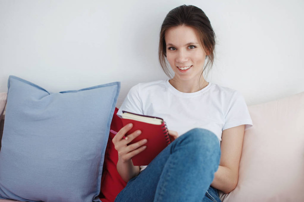 Egy lány, egy fehér pólót és farmert keresztbetett lábbal ül a kanapén párnák szürke, és áttekinti a piros naplója. - Fotó, kép