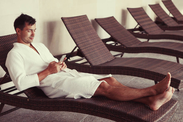 Молодий красивий чоловік лежить на шезлонгу в басейні в білій сукні з террі і дивиться на смартфон
 - Фото, зображення