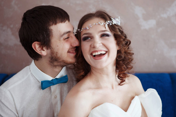 Aranyos menyasszony esküvői fehér fejdísz, haj és szakáll vonzó vőlegény ruha és nyakkendő egy pillangó a kék kanapé, csók és mosolyogva ül. - Fotó, kép