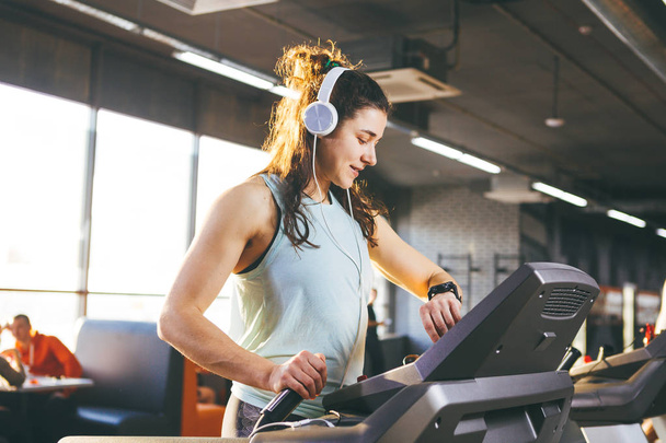 Beautiful young woman with long hair trains in the gym on a treadmill. Listens to music in large headphones. On his arm a sports watch in black for tracking speed, heart rate, pace and stopwatch - Fotografie, Obrázek