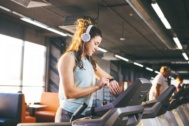 Beautiful young woman with long hair trains in the gym on a treadmill. Listens to music in large headphones. On his arm a sports watch in black for tracking speed, heart rate, pace and stopwatch - Photo, Image
