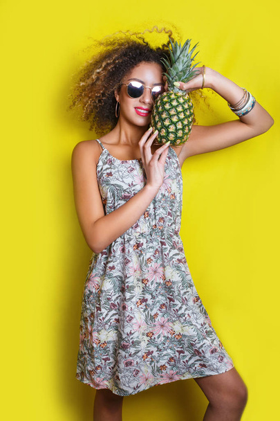 Fashion summer portrait afro american woman in sunglasses and pineapple over yellow background - Fotó, kép