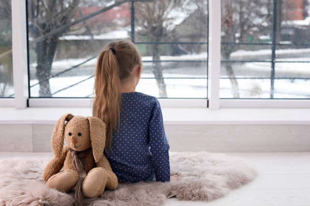 Lonely little girl with toy sitting near window indoors. Autism concept - Fotoğraf, Görsel