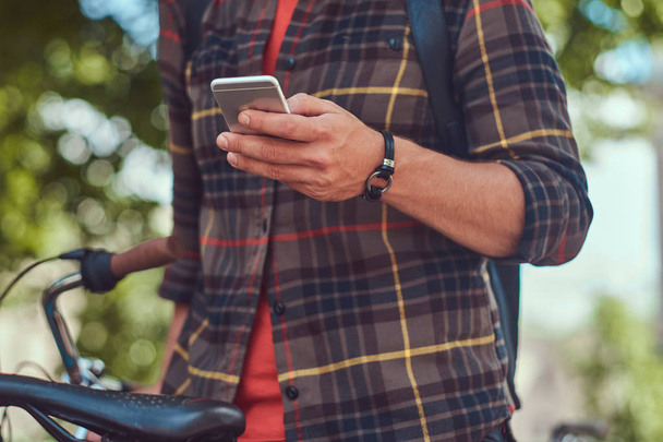 Обрезанное изображение молодой мужчина в шерстяной рубашке, держит смартфон, стоит на стоянке для велосипедов в городском парке
. - Фото, изображение