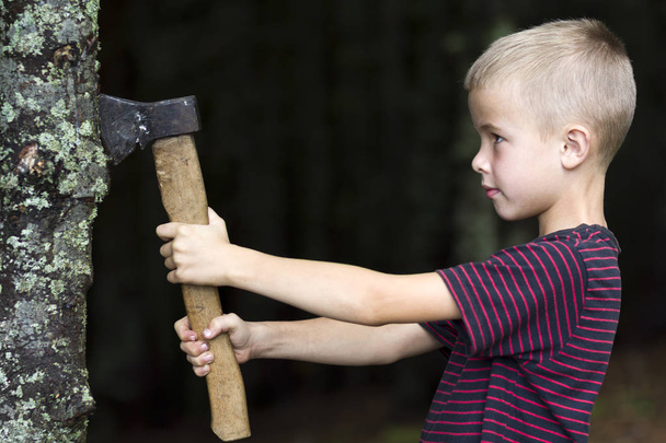 Niño pequeño con hacha pesada de hierro viejo que corta el tronco del árbol en el bosque en el día de verano. Actividades al aire libre y trabajo físico
. - Foto, imagen