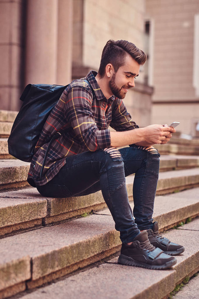 Χαμογελαστά όμορφη μαθητής με μοντέρνο κούρεμα και τα γένια, φορώντας ένα fleece μπλούζα και τζιν, χρησιμοποιώντας ένα smartphone, συνεδρίαση σχετικά με τα βήματα ενάντια σε ένα παλιό κτίριο. - Φωτογραφία, εικόνα
