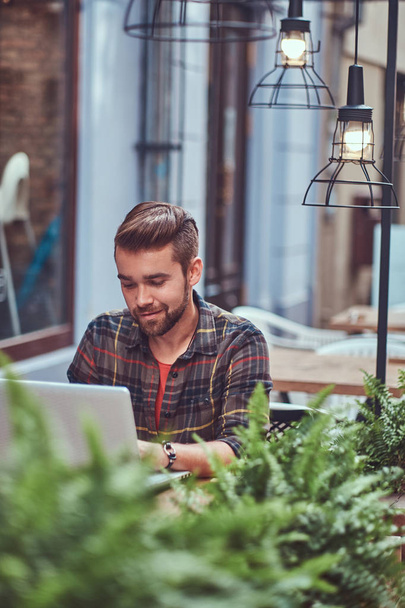 Portret van een jonge mooie freelancer met stijlvolle kapsel en baard, gekleed in een fleece shirt, werken op een laptopcomputer, zittend in een café in de buitenlucht. - Foto, afbeelding