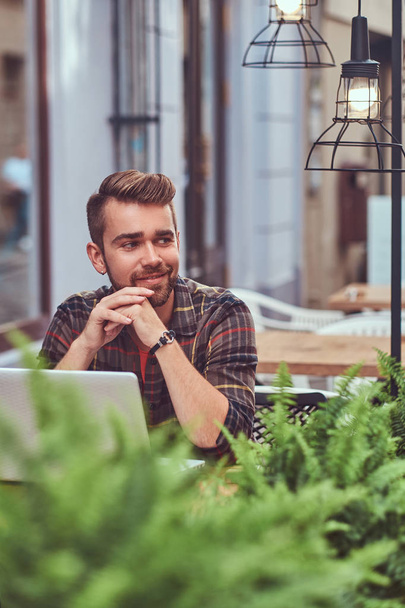 Retrato de um jovem freelancer sorridente com corte de cabelo elegante e barba, vestido com uma camisa de lã, trabalhando em um computador portátil, sentado em um café ao ar livre
. - Foto, Imagem