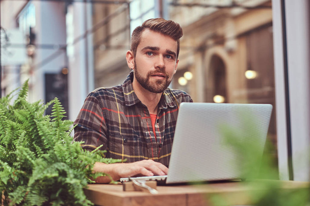 Portret van een jonge mooie freelancer met stijlvolle kapsel en baard, gekleed in een fleece shirt, werken op een laptopcomputer, zittend in een café in de buitenlucht. - Foto, afbeelding