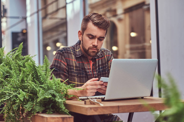 Portret van een jonge freelancer met stijlvolle kapsel en baard, gekleed in een fleece shirt, met behulp van een smartphone tijdens het werken op een laptopcomputer, zittend in een café in de buitenlucht. - Foto, afbeelding