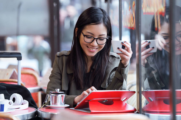 Kaunis nuori aasialainen nainen käyttää hänen digitaalinen tabletti juodessaan kahvia kahvilassa
. - Valokuva, kuva