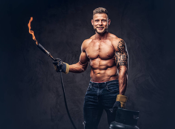 Brutal tattooed male welder with a stylish haircut and beard, with muscular body, dressed in only jeans, holds propane tank and a burning burner, standing in a studio, looking at a camera.  Isolated  - 写真・画像