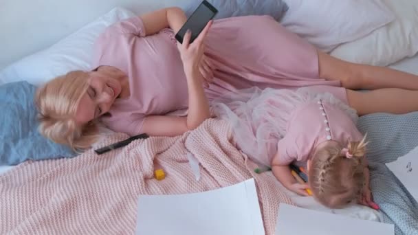 Young attractive blond mom lay on bed with her little charming daughter in pink dresses and mobile phone. - Imágenes, Vídeo