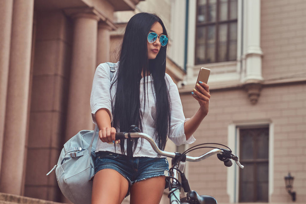 A sexy brunette female wearing blouse and denim shorts in sunglasses, reading the message on a smartphone, standing on steps with a bicycle near an old building in the city. - Foto, Imagen