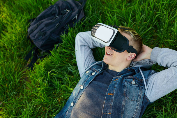 The young man lies on a green grass with VR glasses and relaxing. Outdoor. Copy space.  - Фото, зображення