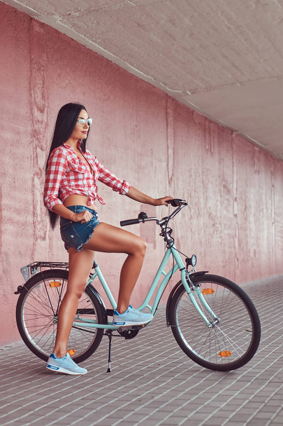 Сексуальна стильна брюнетка дівчина в рожевій фланельній сорочці і джинсових шортах в окулярах, позує з міським велосипедом на рожевій стіні
. - Фото, зображення