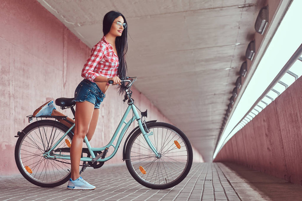 セクシーな笑顔ブルネットの少女、メガネでピンクのフランネルのシャツとデニムのショート パンツを着てピンクの壁に都市の自転車でポーズ. - 写真・画像