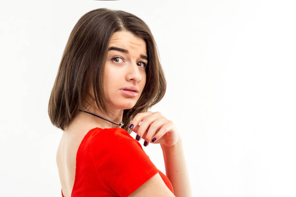 Portrait of a young brunette girl in red t-shirt looking suspiciously over her shoulder into the camera against white background - Φωτογραφία, εικόνα