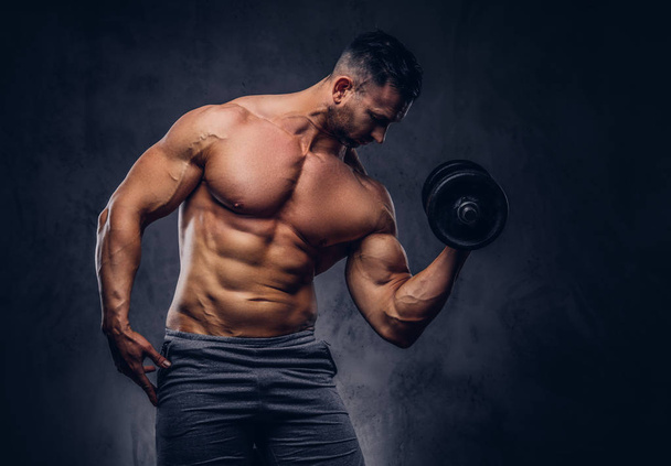 Tall stylish shirtless bodybuilder dressed in sports shorts, doing exercise on a bicep with a barbell, standing in a studio. Isolated on a dark background. - Photo, Image