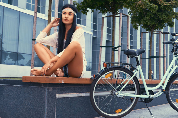 Porträt einer sexy Hippie-Frau in Bluse und Shorts mit Stirnband, sitzt auf einer Bank, kreuzt barfuß die Beine, fährt mit dem Stadtrad gegen einen Wolkenkratzer. - Foto, Bild