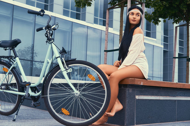 Portrait of a sexy hippie female wearing blouse and shorts in a headband, sits barefoot on a bench near city bike against a skyscraper. - 写真・画像