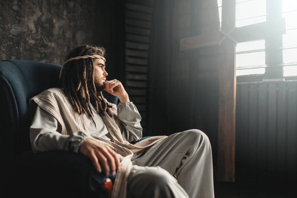 задумчивый Иисус Христос, сидящий в кресле у окна с крестом распятия. Тяжелые времена для христианской веры
 - Фото, изображение
