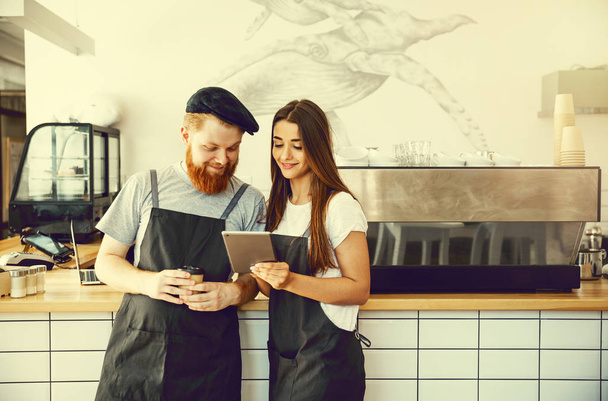 Coffee Business Concept - Positive young bearded man and beautiful attractive lady barista couple in apron looking at camera while standing at bar Counter - Zdjęcie, obraz