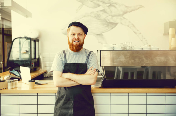 Кофе Бизнес Концепция - Положительный молодой бородатый человек в фартуке смотрит в камеру, стоя у барной стойки
 - Фото, изображение