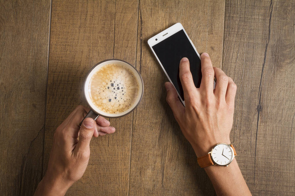 mužské ruce držící smartphone a šálek kávy na dřevěný stůl - Fotografie, Obrázek