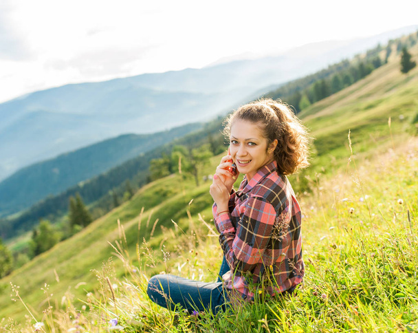 Curly hymyilevä nuori nainen puhuu puhelimeen istuu kaunis maisema vuoren kesällä. Nykyaikaiset ihmiset käyttävät kannettavia mobiililaitteita kaikkialla
. - Valokuva, kuva