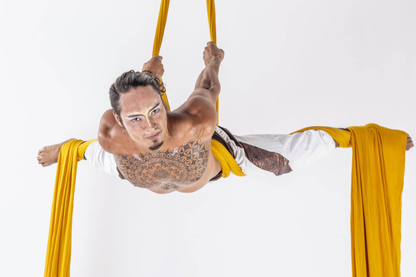 привлекательный мускулистый мужчина, исполняющий акробатические трюки с желтыми тканями на белом фоне
 - Фото, изображение