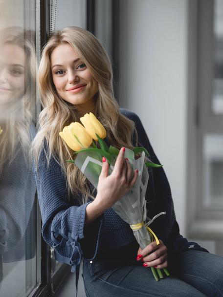 Happy blond woman relaxing with flowers near window - Photo, Image