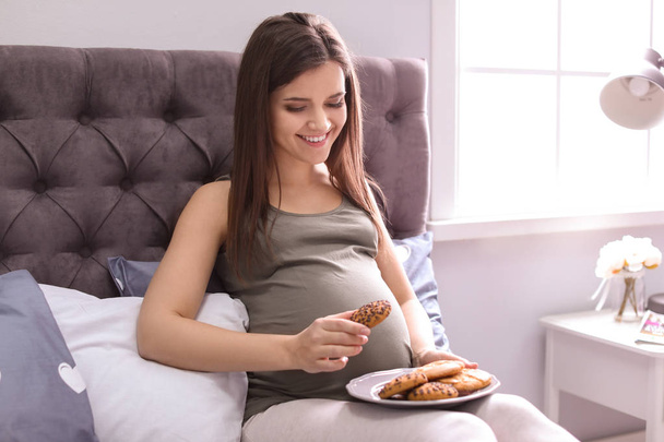 Young pregnant woman holding plate with cookies on bed - Fotó, kép