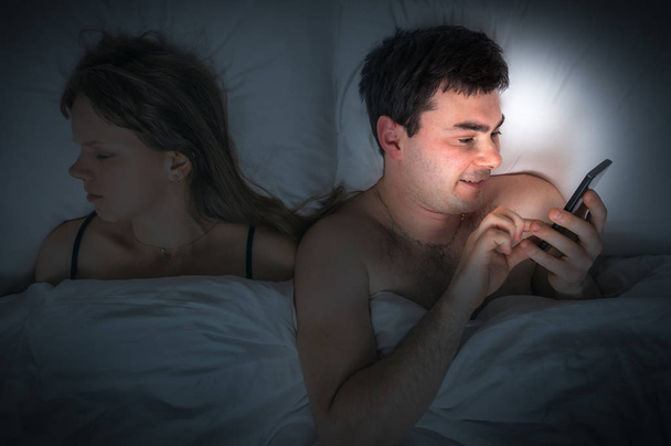 junger Mann mit Handy, während seine Frau nachts schläft - Foto, Bild