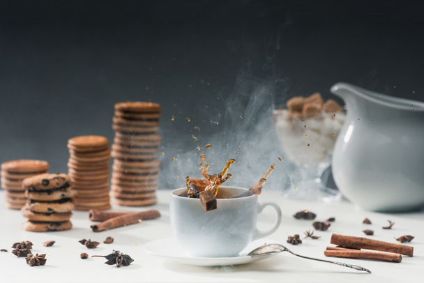 Xícara branca de café quente fumegante com salpicos de cubos de açúcar na mesa com biscoitos e especiarias
 - Foto, Imagem